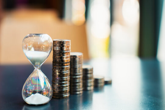 Hourglass with flowing sand and stacked coins on a dark table against a blurred background. Concept of time is money, long-term financial investment, business growth, and wealth.