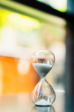 Close up of an hourglass with white sand flowing on a dark table against a bright blurred bokeh background. Concept of time passing, countdown, business deadline, and future planning.