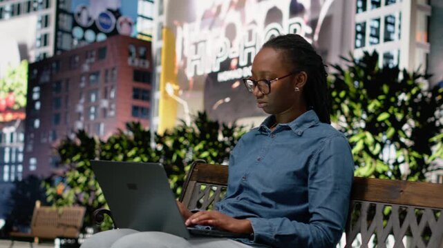 Black woman pleased celebrating successful career win on park bench, feeling joyous and proud with good news. Person in positive mood using laptop and solving tasks outside in city center.