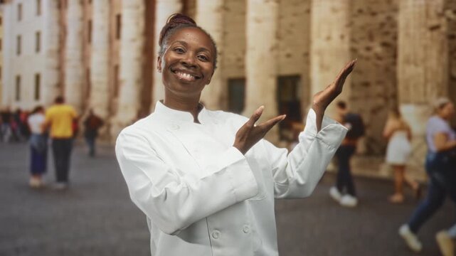 Woman chef in white jacket presenting with hands on a crowded street by stone building, smiling and gesturing to passersby; pride hospitality.