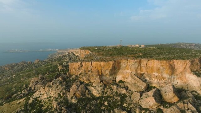 Aerial drone pull-back from rugged limestone cliff plateau on western Malta coastline with sea and haze atmosphere