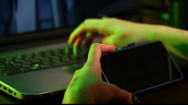 Business woman holding mobile phone in her hands sitting at work table with laptop. Online work, study, research, connection on pc. Closeup workspace in creative art green yellow dark shadow light