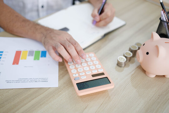 
Man sits at desk, thoughtful, pen in hand. Piggy bank and coins rest nearby, symbolizing savings. He sketches ideas on notebook, surrounded by charts. Office shelves hold colorful folders