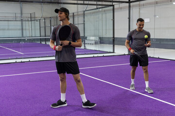 Obraz premium Two male players wearing gray shirts on purple court holding paddles and bouncing yellow ball