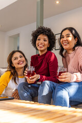 Naklejka premium Diverse female friends sitting on couch in living room holding red drinks near coffee table, tablet