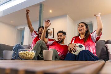 Naklejka premium Friends cheering on gray sofa in living room, wearing red jerseys with soccer ball and popcorn