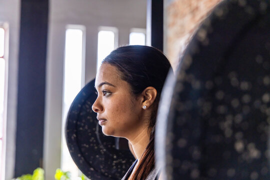 Woman standing in gym facing left, weight plates framing head, windows and brick showing