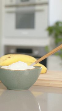 Vertical video: Sliding spoon entering frame, touching bowl on gray counter, making banana snack