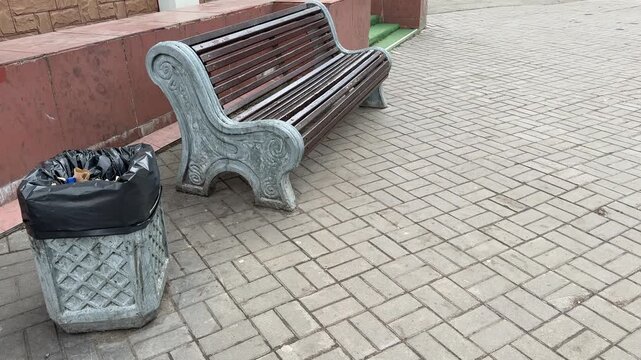 Empty park bench next to trash bin with black bag on gray paving stones during day, decorative cast iron bench ends, red low wall