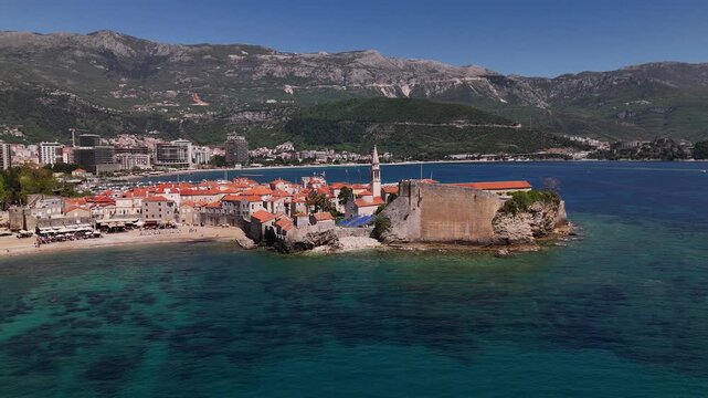 Budva Old Town seen from drone captures medieval stone walls, terracotta roofs, narrow streets and seaside citadel along turquoise Adriatic with mountain backdrop and sandy beaches under clear