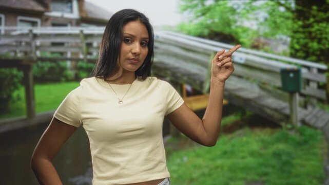 Woman points index finger to right at wooden bridge in forest wearing cream t shirt and gold necklace; confidence assertiveness guidance.