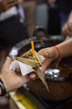 mano de mujer entregando un tamal con mole verde y mole rojo, mano de hombre recibiendo, sobre una olla de mole