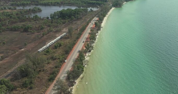 Backward aerial view shows a train traveling on tracks parallel to a coastal road, separated by trees from the turquoise sea.