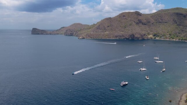 Aerial footage of Soufriere Bay in Saint Lucia featuring colorful Caribbean town nestled between lush green Piton mountains with sailboats and speedboat crossing turquoise waters.