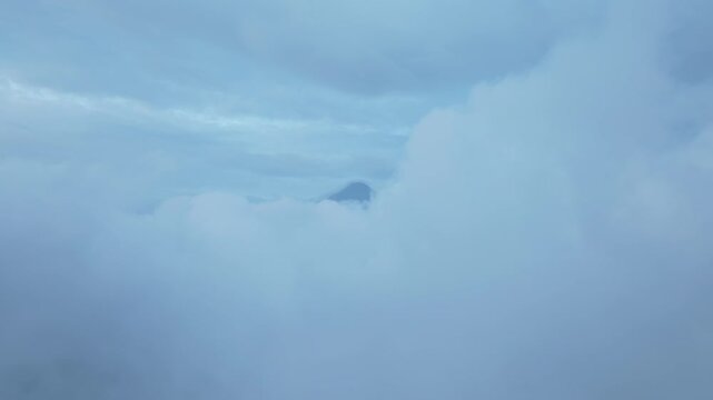 Dramatic mountain summit breaks through thick layers of clouds and fog creating an ethereal misty landscape. Soft blue tones and swirling cloud formations surround the distant peak.