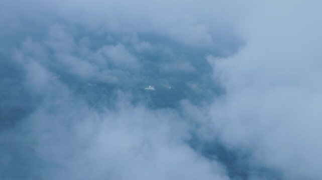 Aerial footage flying above thick white clouds with glimpses of lush green forested terrain visible through gaps in the cloud cover. Soft blue atmospheric haze creates dreamy mood.