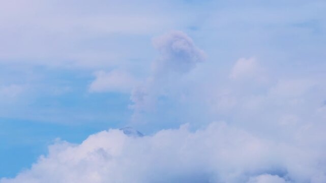 Majestic volcanic eruption in Guatemala as Fuego volcano emits towering gray ash plume rising dramatically through layers of soft white clouds against a serene pale blue sky during daytime.