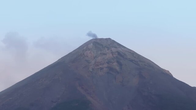 Active Fuego Volcano in Guatemala releases dark smoke plume from its rugged summit. The conical mountain dominates the frame against pale blue sky with wispy clouds drifting past the volcanic peak.