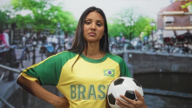 Woman young brunette in brazil t shirt points finger to chest while holding soccer ball on a canal street bridge with bicycles; pride team support.