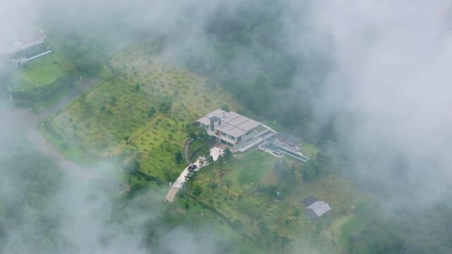 Drone footage reveals modern luxury mansion with swimming pool nestled in lush green landscape. Thick fog and clouds drift across the rural estate creating a mystical atmosphere.