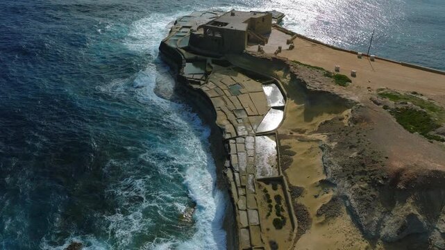 Cinematic drone flyover of Qolla l-Bajda salt pans on Gozo coastline Malta. shot follows a narrow limestone coastal strip extending into Mediterranean Sea, where traditional salt basins 