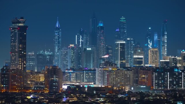 Stunning nighttime aerial view of Dubai Marina skyline featuring illuminated skyscrapers with glowing windows against deep blue twilight sky, residential areas in foreground creating urban layers.