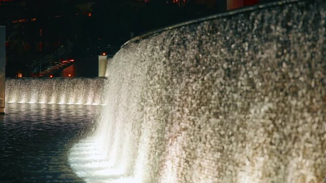 Dubai decorative waterfall fountain illuminated with warm underwater lights at night. Water cascades down curved stone wall into reflecting pool with city lights glowing in background.