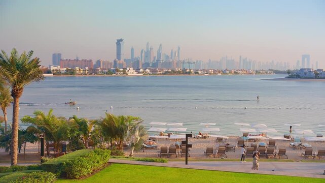 Dubai luxury beach resort with sun loungers and umbrellas on sandy shore. Tourists relax while jet ski passes calm waters. Palm trees frame the Dubai Marina skyline in golden morning haze.