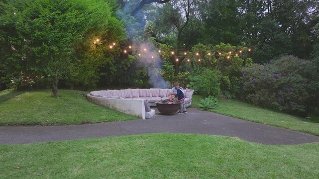 Father and young son spend quality time near smoking fire pit in beautifully landscaped backyard garden with curved sectional sofa seating area, decorative string lights hanging overhead.