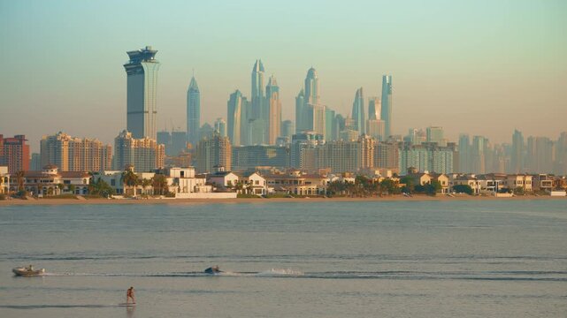 Dubai skyline towers above Palm Jumeirah villas as jet skis and boats cruise through calm turquoise waters during golden sunset. Modern skyscrapers create stunning backdrop for water recreation.