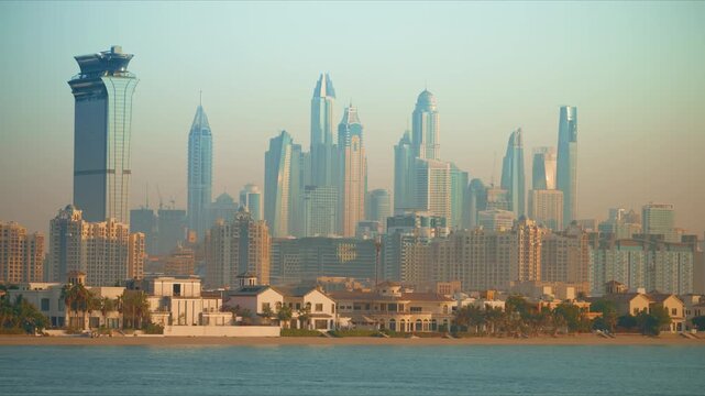 Dubai cityscape featuring iconic Marina skyline with gleaming glass skyscrapers rising behind luxury beachfront villas. Calm turquoise waters reflect hazy golden light as palm trees line sandy shore.
