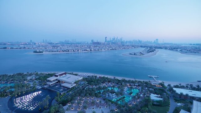 Dubai luxury hotel room balcony overlooking Palm Jumeirah artificial island with resort pools and beach at twilight. City skyline silhouette visible in hazy blue hour light with calm waters below.