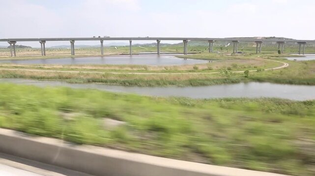 야외 수변 생태 공원 고가 교량 주행 전경 자재: 맑은 날 도로 가드레일 위로 전경의 풀밭이 빠르게 스쳐 지나가는 가운데 중앙의 넓은 강물과 후경을 수평으로 길게 가로지르는 고가도로 교각 구조물을 포착한 패닝 와이드 숏 영상입니다. 자연 경관을 담았으며 교통 인프라, 주행 스케치, 교량 건축물 및 여행 콘셉트의 생생한 풍경 인서트 숏을 상징합니다.