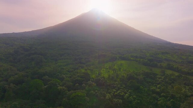 Aerial drone footage captures a majestic volcanic cone rising above lush green tropical rainforest at golden hour, with soft sunlight creating lens flares and a warm hazy pink sky atmosphere.