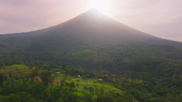 Aerial drone footage captures the majestic Arenal Volcano at sunrise in Costa Rica. Sun rays illuminate the lush green rainforest and small rural houses nestled in the tropical landscape below.