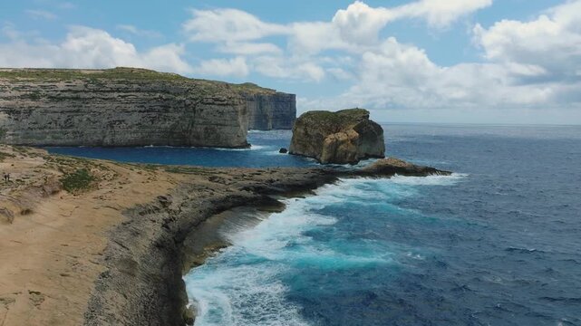 Cinematic drone approach to Fungus Rock sea stack at Dwejra coast on Gozo Malta.  The shot reveals dramatic coastal geology with rugged cliffs and rock formations rising from the Mediterranean Sea