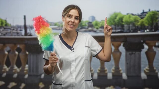 Woman holding a rainbow feather duster pointing index finger on a bridge overlooking river and balustrade; cheerful service.