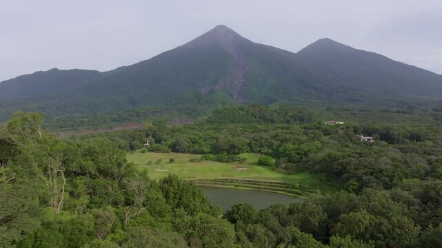 Drone footage reveals scenic golf course nestled among dense tropical forest with majestic volcanic peaks rising in the background under overcast skies and serene pond in foreground.