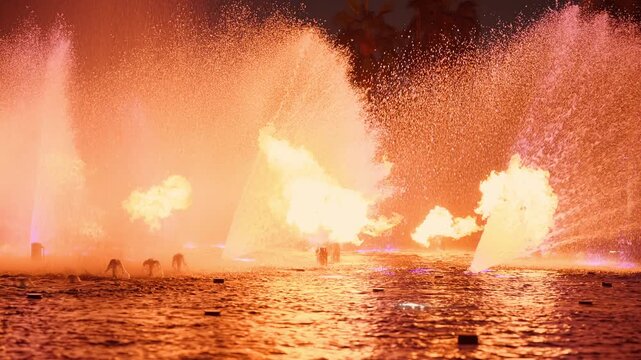 Dubai fountain show features dramatic fire bursts erupting through illuminated purple water jets at night. Palm trees silhouette against glowing mist as flames reflect on dark water surface.