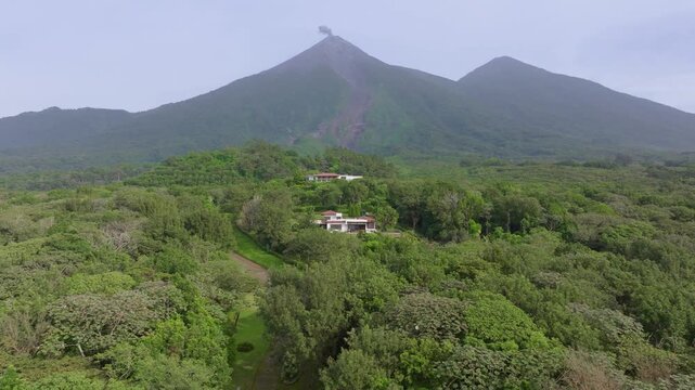 Aerial drone footage captures the active Fuego Volcano emitting smoke above dense tropical rainforest in Guatemala. Lush green canopy surrounds rural homes nestled in the volcanic highlands landscape.