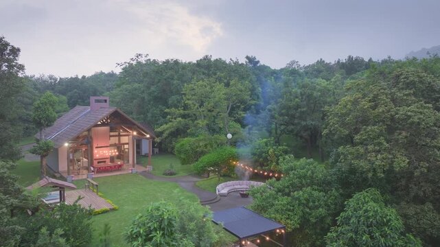 Father and son spending quality time by bonfire in backyard of their house. Aerial view of cozy cabin surrounded by lush green forest with warm lights glowing at dusk and smoke rising from fire pit.