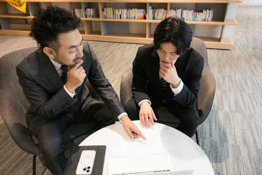 本棚のある社長室で資料を見ながら相談、ビジネスマンの打ち合わせ　契約書など行政書士や士業のイメージ困る考える悩む俯瞰のイメージ