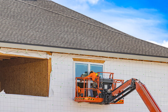 A wide horizontal view of construction workers in an orange boom lift basket installing a window on a multi-story residential building wrapped in housewrap beneath a completed shingle roof, USA