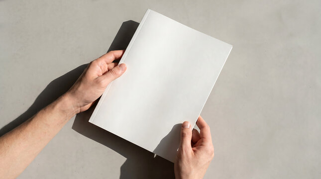 Top View of Hands Holding Blank White Magazine Book Cover Mockup on Concrete Surface with Hard Shadows
