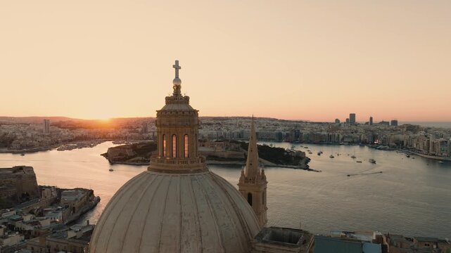 Cinematic aerial drone pullback from dome of Basilica of Our Lady of Mount Carmel in Valletta, Malta with sunset sun briefly hidden and reappearing behind church silhouette