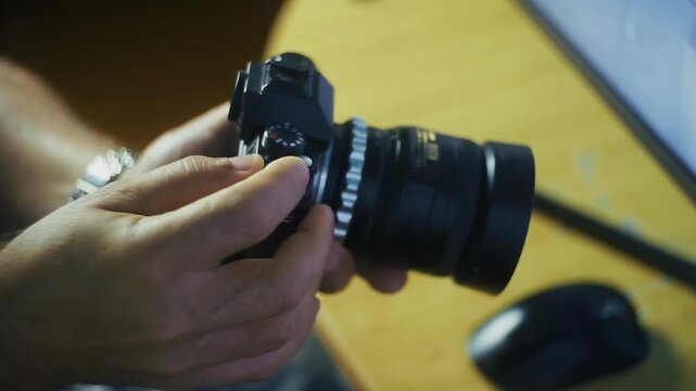 A pair of hands holds a camera with an attached lens and controls it. A computer mouse is visible on the table in the background.
