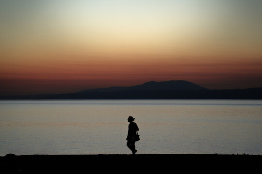Silueta de un joven con el pelo recogido y una bolsa al hombro caminando al atardecer por la orilla de la playa.