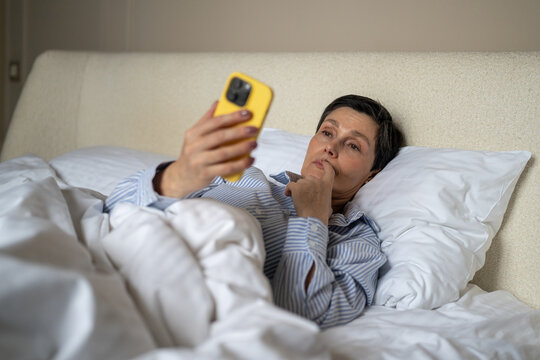 Thoughtful elderly woman in bed procrastinating watching news online on smartphone after waking up in morning, older female reading social media on phone, unhealthy habit using cellphone after sleep.