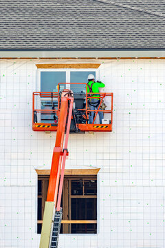 A vertical full shot of a construction worker wearing a safety harness and high-visibility shirt inside a boom lift basket, installing a window on a multi-story residential building wrapped in housewr