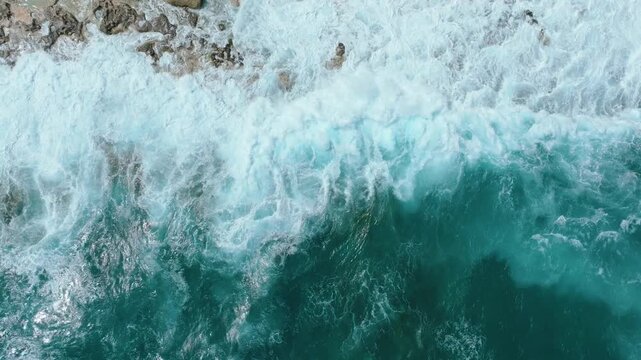 Top down drone view of ocean waves crashing on rocky coastline in Malta with sea foam and textured shoreline in slow motion. Powerful waves roll toward a rocky shoreline and break against the coast